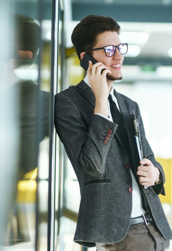 Young businessman in glasses talking on cell phone standing in the modern office.